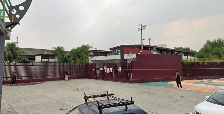 Fotografía en la que se muestra una explanada que da acceso al plantel. Al fondo de la explanada se encuentra la entrada del plantel, conformada por rejas de color rojo que rodean las instalaciones.